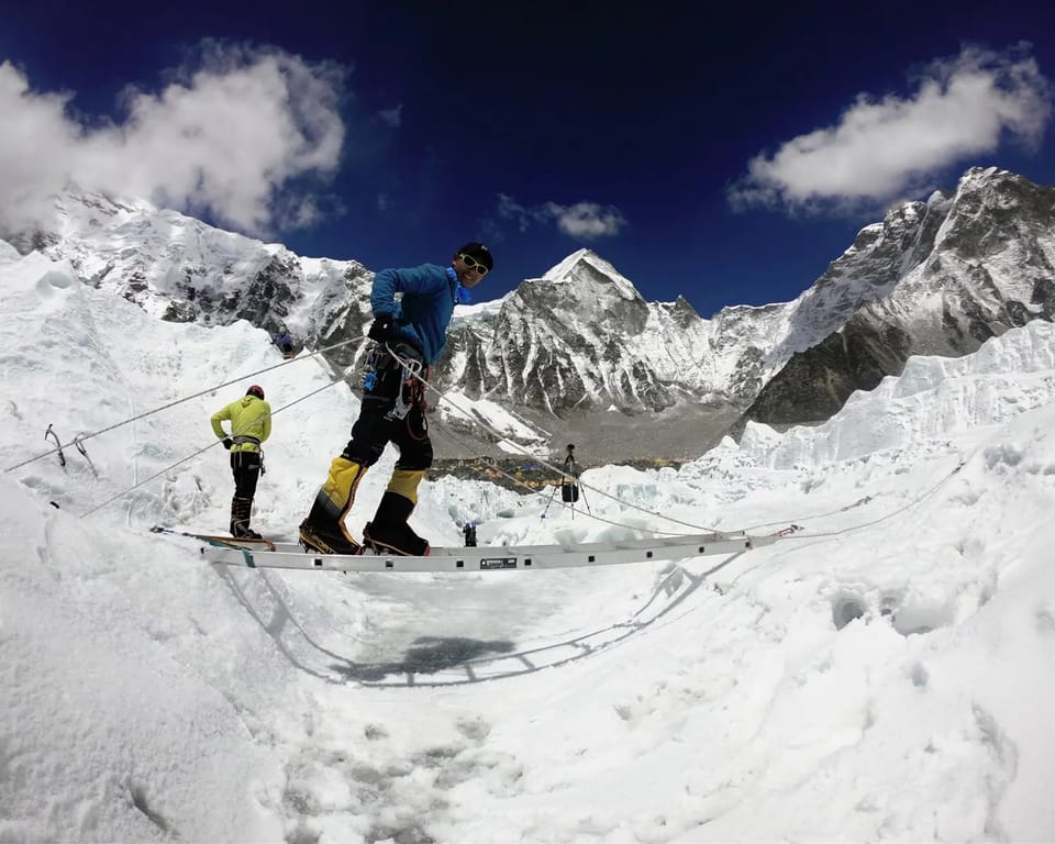 A climber in a blue jacket standing on an aluminium ladder across a crevasse, clipped to fixed ropes, with a tented camp below A climber in a blue jacket standing on an aluminium ladder across a crevasse, clipped to fixed ropes, with a tented camp below